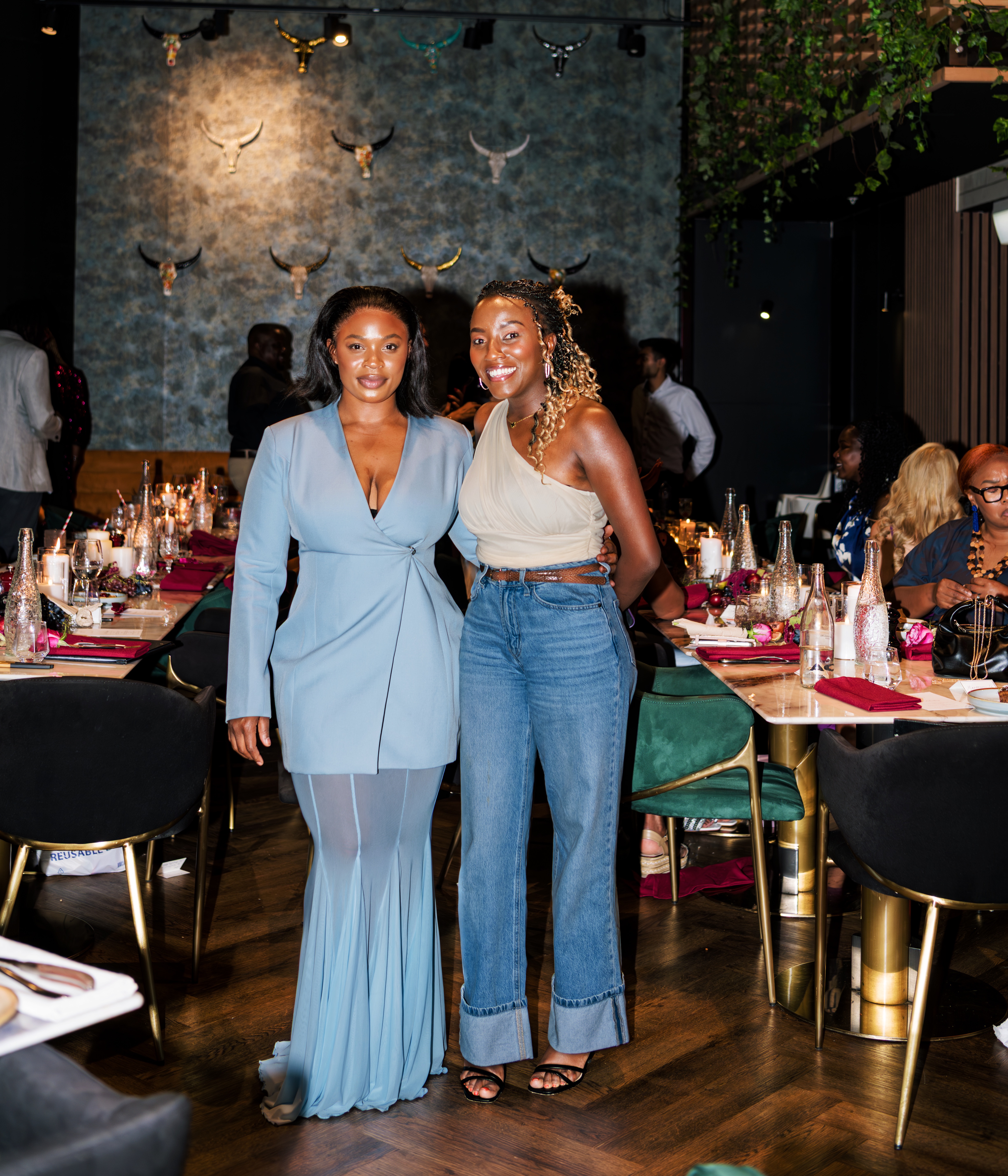 Two women standing together at an elegantly set dining event, surrounded by decorated tables with candles, glassware, and floral accents, in a warmly lit, upscale venue.