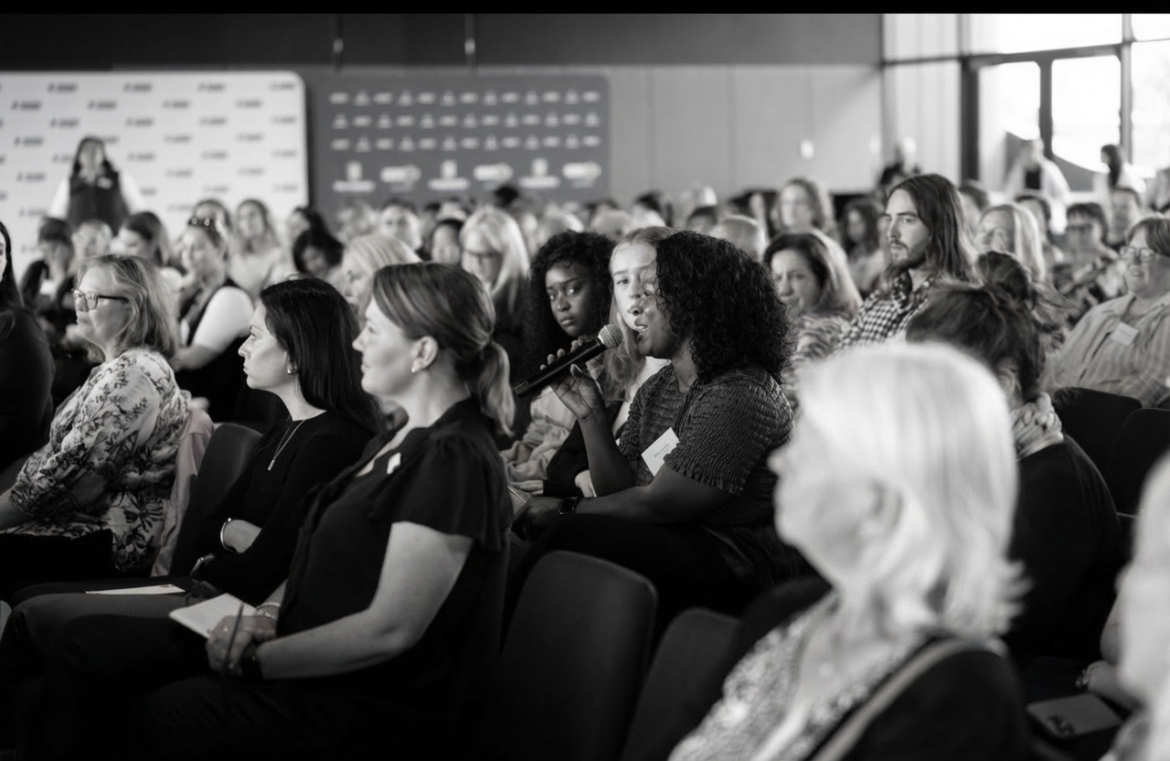 A black woman (Saran Konteh) speaking into a microphone during a large audience event, surrounded by white attendees listening attentively in a conference setting.